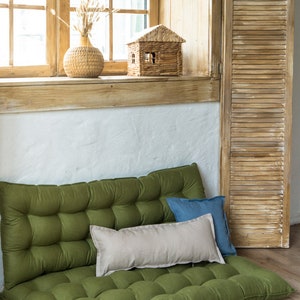May include: A green, tufted floor cushion with two pillows, one beige and one blue, placed on top. The cushion is in front of a window with wooden blinds.