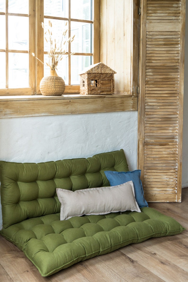 May include: A green, tufted floor cushion with two pillows, one beige and one blue, placed on top. The cushion is in front of a window with wooden blinds.