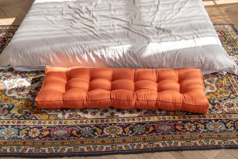 K&ouml;nnte beinhalten: Ein rechteckiges, hellorangefarbenes Kissen mit Kn&ouml;pfen. Das Kissen liegt auf einem gemusterten Teppich.