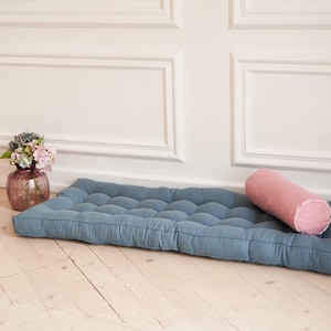 K&ouml;nnte beinhalten: Ein blaues, gestepptes Bodenkissen mit einem rosa, zylindrischen Kissen darauf. Das Kissen liegt auf einem hellen Holzboden vor einer wei&szlig;en Wand mit dekorativen Leisten.