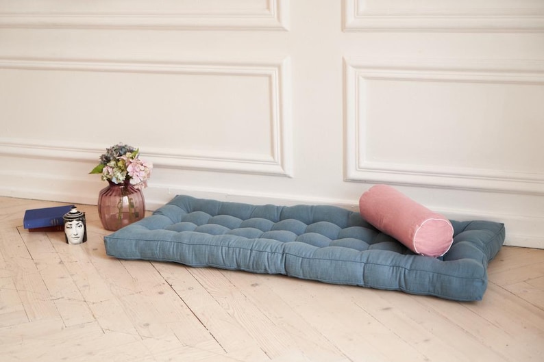 K&ouml;nnte beinhalten: Ein blaues, gestepptes Bodenkissen mit einem rosa, zylindrischen Kissen darauf. Das Kissen liegt auf einem hellen Holzboden vor einer wei&szlig;en Wand mit dekorativen Leisten.