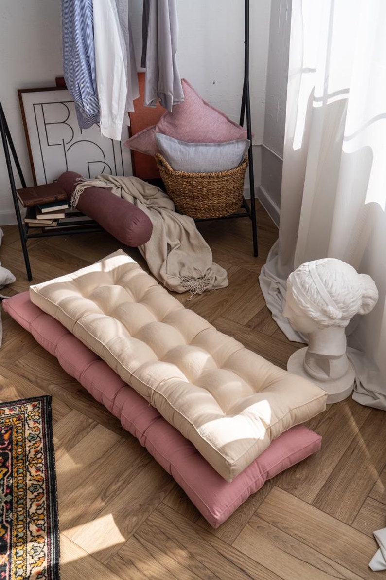 K&ouml;nnte beinhalten: Zwei Bodenkissen, eines beige und eines rosa, liegen &uuml;bereinander auf einem Holzboden. Das beige Kissen hat ein gestepptes Design und das rosa Kissen ist einfarbig.