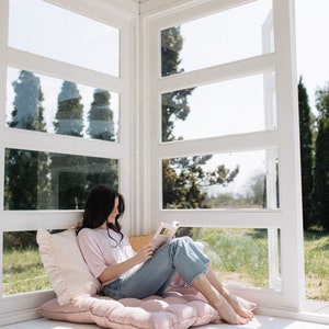 May include: A woman sits on a pink cushion in a sunroom, reading a book. The room has large windows with white trim and a white ceiling.