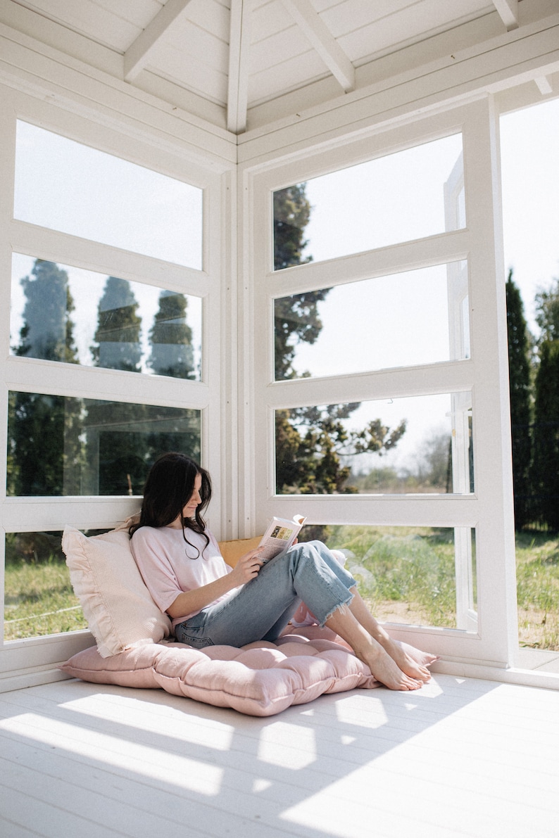May include: A woman sits on a pink cushion in a sunroom, reading a book. The room has large windows with white trim and a white ceiling.