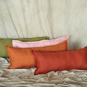 Peut inclure: Quatre coussins rectangulaires en lin dans les tons de vert, rose, orange et rouge-orange sont disposés sur une surface texturée de couleur neutre. Les coussins ont un design simple et le tissu semble légèrement froissé.