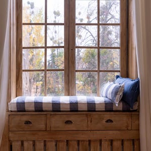 May include: A window seat with a white and blue striped cushion and two pillows. The window is open and there are trees visible outside. The seat is built into a wooden frame with drawers underneath.