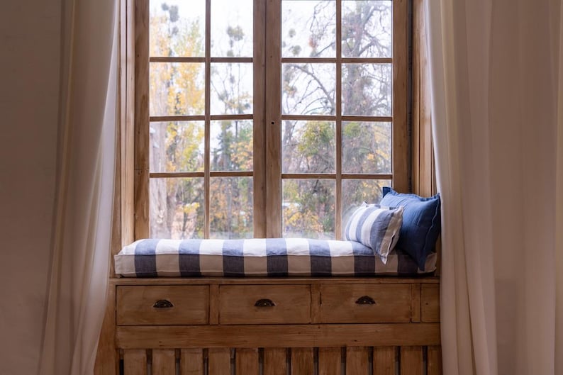 May include: A window seat with a white and blue striped cushion and two pillows. The window is open and there are trees visible outside. The seat is built into a wooden frame with drawers underneath.