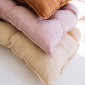 May include: Three linen cushions stacked on top of each other. The top cushion is brown, the middle cushion is pink, and the bottom cushion is beige.