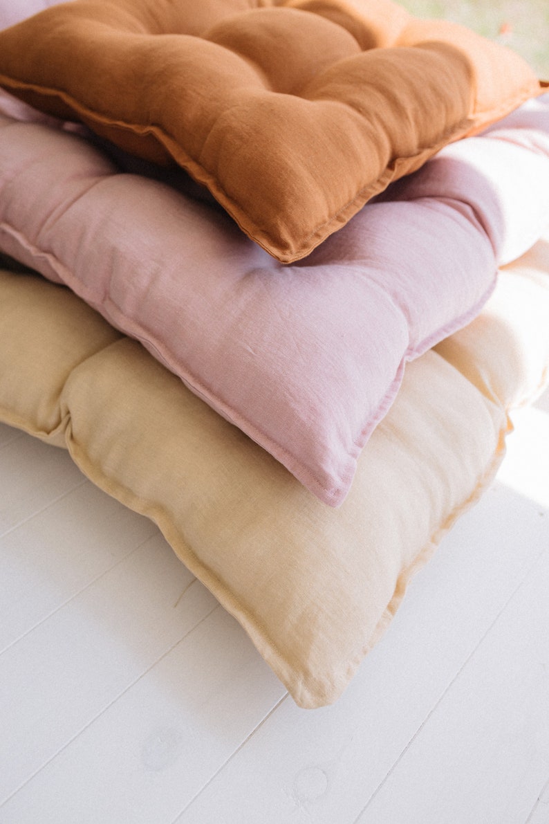 May include: Three linen cushions stacked on top of each other. The top cushion is brown, the middle cushion is pink, and the bottom cushion is beige.