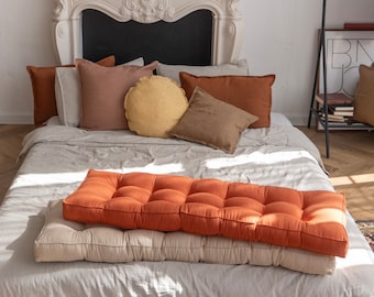 May include: A bed with a white linen duvet cover and a variety of pillows in shades of brown, beige, and yellow. The bed has a thick, orange, tufted cushion on top of the duvet.