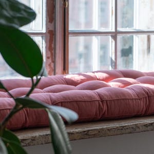 K&ouml;nnte beinhalten: Ein rosa gestepptes Kissen auf einer Fensterbank. Das Kissen ist aus einem weichen, strukturierten Stoff und hat einen leicht abgenutzten Look.