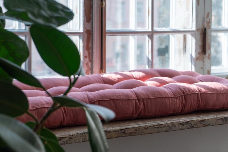K&ouml;nnte beinhalten: Ein rosa gestepptes Kissen auf einer Fensterbank. Das Kissen ist aus einem weichen, strukturierten Stoff und hat einen leicht abgenutzten Look.