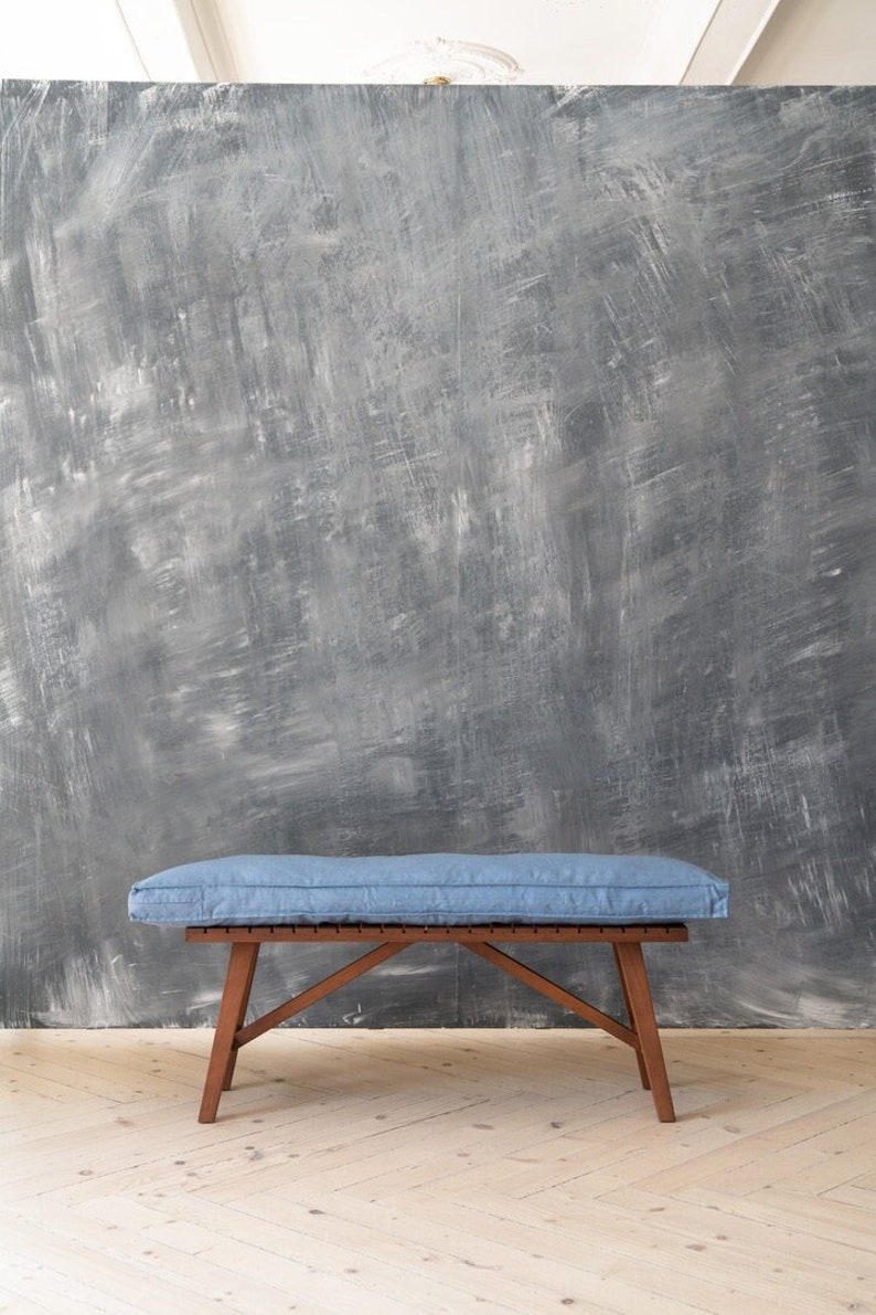 Op de afbeelding: Een houten bank met een blauw kussen. De bank heeft een eenvoudig ontwerp met vier poten en een lattenbodem. De bank staat voor een grijze, getextureerde muur.
