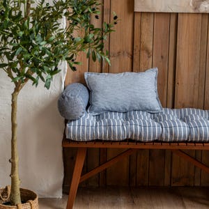 Op de afbeelding: Een houten bank met een blauw-wit gestreept kussen en een blauw cilindrisch kussen. De bank staat tegen een houten muur en op de voorgrond staat een plant in een pot.