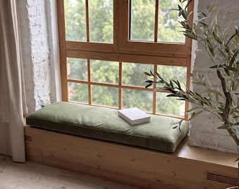 Pode incluir: Uma almofada retangular verde-sálvia está sobre um banco de madeira junto à janela. A almofada é lisa e plana, com bordas debruadas. Um livro branco fechado repousa no centro da almofada, em frente a uma janela grande.