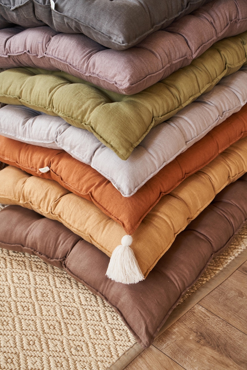 May include: A stack of seven floor cushions in various shades of brown, beige, and green. The cushions are made of a soft, textured fabric and have a tufted design. The cushions are arranged in a staggered pattern, with the top cushion slightly offset from the bottom cushion. The cushions are sitting on a woven rug.