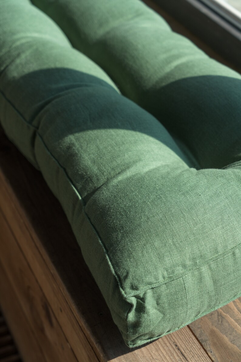 K&ouml;nnte beinhalten: Ein gr&uuml;nes Leinenkissen mit gestepptem Design. Das Kissen liegt auf einer Holzoberfl&auml;che.