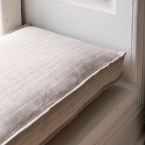 May include: A close-up of a beige striped cushion with a white border. The cushion is placed on a white window seat.