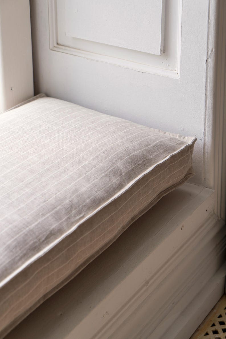 May include: A close-up of a beige striped cushion with a white border. The cushion is placed on a white window seat.