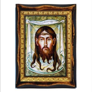May include: A framed icon depicting the face of Jesus Christ. The icon is painted in a traditional style with a gold background and a green and white patterned cloth behind the face. The icon is framed in a dark wood frame with a gold leaf border.