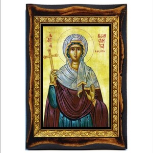 Può includere: Un'icona religiosa incorniciata che raffigura una donna che tiene una croce. L'icona è dipinta in uno stile tradizionale con colori vivaci e dettagli intricati. La cornice è in legno con finitura in foglia d'oro e presenta un bordo decorativo di foglie.