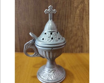 May include: A silver metal censer with a cross on top and intricate floral designs. The censer has a hinged lid and a handle.