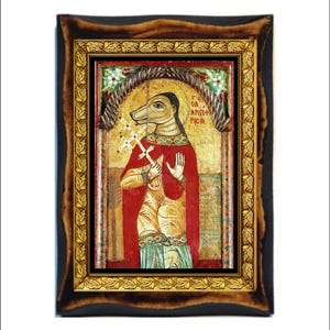 May include: Framed religious icon featuring a figure with a dog's head, wearing a red robe and holding a cross. The icon has gold accents and is set in a dark wooden frame with a gold inner border. The background is gold with faded text.