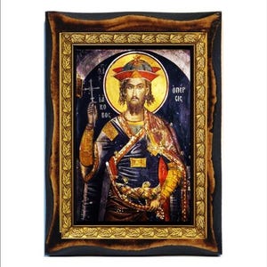 Puede incluir: Icono de una figura religiosa en un marco de madera oscura con un borde interior dorado. La figura tiene un halo dorado y viste un sombrero rojo y ropa ornamentada. El fondo es oscuro con texto griego.