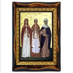 May include: A framed icon depicting three women in traditional clothing, with a gold leaf border and a black wooden frame. The icon is titled "Ai Dias Tres Moteres" in Greek.