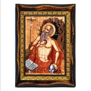 May include: A religious icon depicting a bearded man holding a skull and a book. The man is wearing a red robe and is sitting in a cave. The icon is framed in a dark wood frame with gold accents.