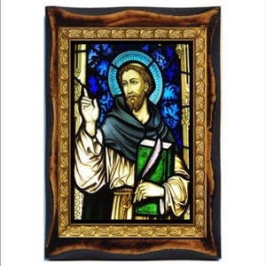 May include: A stained glass window depicting a man in a brown robe holding a book. The window is framed in a dark wood frame with a gold leaf border.