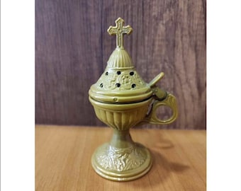 May include: A gold-colored metal incense burner with a cross on top and intricate floral designs. The burner has a handle and sits on a wooden surface.