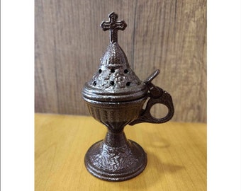 May include: A small, ornate, dark brown metal incense burner with a cross on top. The burner has a round base and a handle on the side.