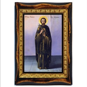 Puede incluir: Un icono enmarcado de San Antonio del Desierto, una figura religiosa de la fe cristiana. El icono está pintado en un estilo tradicional con un marco dorado y un fondo de madera oscura. El icono representa a San Antonio de pie con una cruz y un rosario en sus manos. El texto en el icono dice "O' Agios Antonios" y "The Hermit".