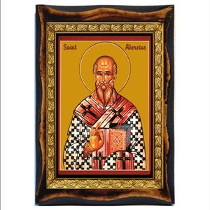 May include: A framed icon of Saint Abercius, a religious figure depicted in a red and white checkered robe with a gold halo. The icon is set against a gold background with a red border and a gold frame.