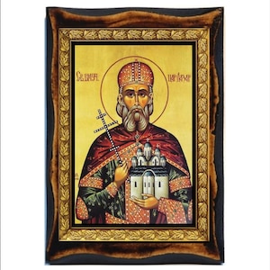 May include: An icon featuring a religious figure with a halo, holding a cross and a miniature church. The figure is dressed in ornate robes with gold and red accents. The icon is framed in a dark wood frame with a gold leaf border.