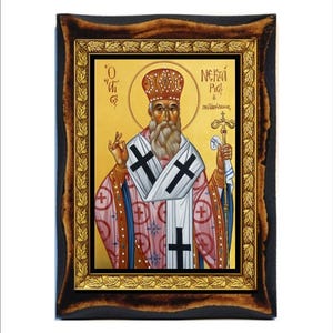 May include: An icon of a religious figure in a gold frame. The figure has a halo, a long white beard, and is wearing a red and white robe with a cross design. The frame is dark brown with gold accents.