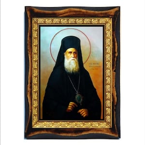 May include: Framed religious icon featuring a portrait of a man with a long white beard, wearing a black robe and hat. The icon has a gold border and a dark brown wooden frame. The background includes a halo.