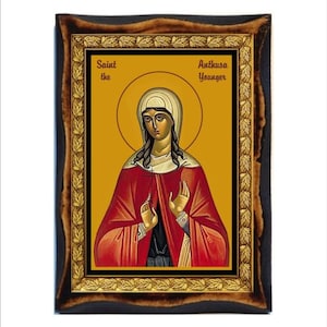 May include: A framed icon of Saint Anthusa the Younger. The icon is painted in a traditional style with a gold background and a red robe. The saint is depicted with a cross in her hands.