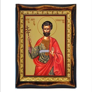 Könnte beinhalten: Ikone eines Mannes mit Heiligenschein, der ein Kreuz hält, in einem goldgerahmten Bild. Der Mann trägt ein rotes Gewand und eine grün-goldene Tunika. Der Rahmen ist schwarz mit einem goldenen Innenrand und einem dekorativen Blättermuster.