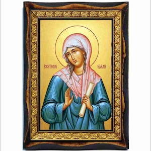 May include: A rectangular religious icon featuring a painting of Righteous Sarah with a gold halo, wearing a pink head covering and blue robe, holding a scroll. The image is set within a decorative gold frame with leaf patterns and a dark border.