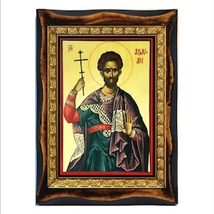 Può includere: Un'icona incorniciata di un santo che tiene una croce. L'icona è dipinta in uno stile tradizionale con uno sfondo dorato e una cornice in legno scuro. Il santo indossa una tunica verde e una fascia rossa. Il testo "СВ. АЛЕКСАНДР" è scritto in caratteri cirillici nella parte superiore dell'icona.