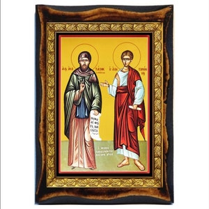 May include: A framed icon depicting two figures, one in green and red robes, the other in red and white robes. The figures are holding scrolls and are standing on a yellow background. The frame is brown with gold accents and a leaf design.