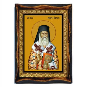 May include: Icon of a religious figure with a long white beard, wearing a black hat and ornate robes. The icon is set against a gold background and framed in a dark wood frame with gold accents. The text "ΑΓΙΟΣ ΝΕΚΤΑΡΙΟΣ" is at the top.