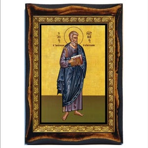 Puede incluir: Un icono de una figura religiosa, enmarcado en un marco de madera oscura con un borde interior dorado. La figura está representada con túnicas azules y moradas, sosteniendo un libro. El fondo es dorado con texto griego.