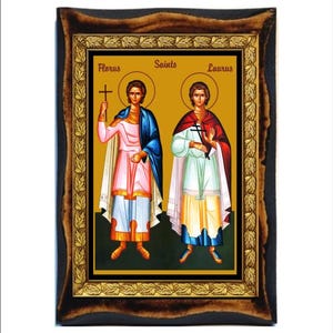 Può includere: Un'icona incorniciata che raffigura due santi, Florus e Laurus, con uno sfondo dorato e un bordo decorativo. I santi sono raffigurati in stile bizantino tradizionale, con aureole e abiti decorati.