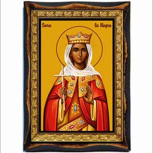 May include: A rectangular wooden plaque featuring a religious icon of Sara la Negra. The figure wears a gold crown, a white head covering, and a red robe adorned with colourful jewels, holding a cross. The frame has a gold leaf pattern.