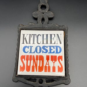 May include: Black cast iron trivet with a white tile insert. The tile reads "KITCHEN CLOSED SUNDAYS" in black, blue, and red lettering. The trivet has a decorative handle and a rectangular shape.