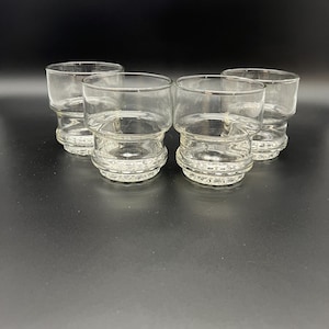May include: Four clear, vintage-style drinking glasses. Each glass has a thick, textured base and a cylindrical upper portion. The glasses are made of transparent glass, allowing visibility of the contents. The glasses are arranged on a dark surface.