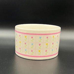 May include: A cylindrical ceramic container with a cream-colored base. The container is decorated with a pink band at the top and bottom, and a pattern of pink, yellow, and blue tulips on a white grid.